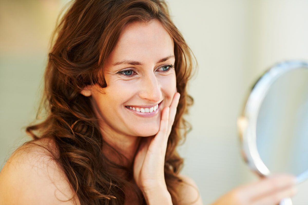 woman with long brown hair looking to a hand mirror and smiling at herself